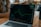 A laptop displaying a line graph with various peaks and troughs on its screen, indicating stock or financial data. The graph and numbers are green against a black background. The laptop is placed on a wooden table, and beside it, a person's arm is partially visible.