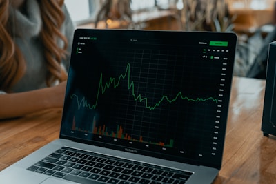 A laptop displaying a line graph with various peaks and troughs on its screen, indicating stock or financial data. The graph and numbers are green against a black background. The laptop is placed on a wooden table, and beside it, a person's arm is partially visible.