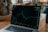A laptop displaying a line graph with various peaks and troughs on its screen, indicating stock or financial data. The graph and numbers are green against a black background. The laptop is placed on a wooden table, and beside it, a person's arm is partially visible.