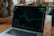 A laptop displaying a line graph with various peaks and troughs on its screen, indicating stock or financial data. The graph and numbers are green against a black background. The laptop is placed on a wooden table, and beside it, a person's arm is partially visible.