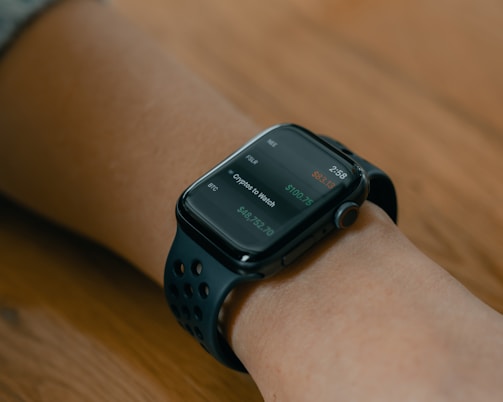 A smartwatch with a dark screen displaying cryptocurrency prices on a person's wrist. The watch has a modern design with a smooth black strap featuring circular cutouts. The background is a wooden surface, adding warmth to the composition.