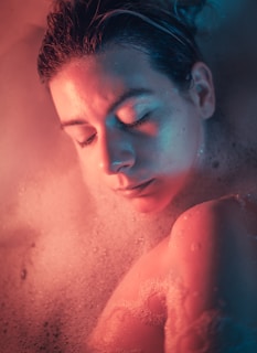 A person with closed eyes is lying in a bath filled with foam. The image is illuminated by a soft, ethereal mix of pink and blue lighting, creating a serene atmosphere.
