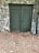 A stone wall features a pair of green double doors with louvered panels. The ground is covered with dry pine needles and scattered leaves. A metal downspout is attached at one side of the wall.
