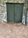 A stone wall features a pair of green double doors with louvered panels. The ground is covered with dry pine needles and scattered leaves. A metal downspout is attached at one side of the wall.