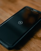 A smartphone lies on a wooden surface, displaying a logo resembling a 'W' inside a circle on its screen. The phone is encased in a protective cover.
