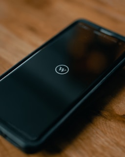 A smartphone lies on a wooden surface, displaying a logo resembling a 'W' inside a circle on its screen. The phone is encased in a protective cover.