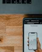 A close-up view of a smartphone displaying a financial app with a graph and numerical data, held by a hand. The screen shows the brand 'Wealthsimple'. Above it is part of a laptop keyboard with keys visible. The background consists of a wooden surface.