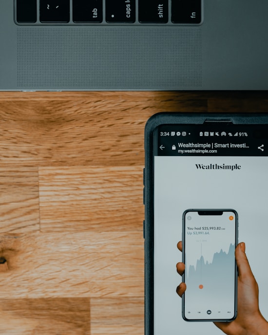 A close-up view of a smartphone displaying a financial app with a graph and numerical data, held by a hand. The screen shows the brand 'Wealthsimple'. Above it is part of a laptop keyboard with keys visible. The background consists of a wooden surface.