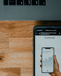 A close-up view of a smartphone displaying a financial app with a graph and numerical data, held by a hand. The screen shows the brand 'Wealthsimple'. Above it is part of a laptop keyboard with keys visible. The background consists of a wooden surface.
