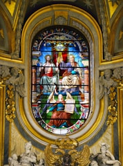 A detailed stained glass window depicts sacred figures, including two seated individuals draped in elaborate robes, one holding a sphere, and the other appearing in a blessing gesture. An angel kneels below them, holding a tablet with writing. The background is rich with vibrant colors and ornate patterns, surrounded by intricate architectural details and sculptures.