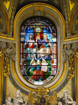 A detailed stained glass window depicts sacred figures, including two seated individuals draped in elaborate robes, one holding a sphere, and the other appearing in a blessing gesture. An angel kneels below them, holding a tablet with writing. The background is rich with vibrant colors and ornate patterns, surrounded by intricate architectural details and sculptures.