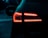 A close-up view of a car's rear lights shining brightly in the dark, with red LED patterns. The car surface is wet, likely due to rain, and the background is dimly lit.