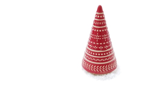 A cone-shaped, red ceramic object with intricate white patterns on its surface, resembling decorative elements or leaves. It sits on a plain white background, suggesting snow or a wintry setting.
