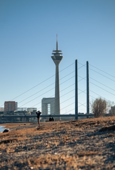 A tall telecommunications tower stands prominently against a clear sky. The scene includes a modern bridge with cables and buildings in the background. In the foreground, a person is taking a photograph, while another is sitting on the ground.