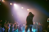 A person with long hair, wearing a dark top and light pants, is performing on stage under bright spotlights. In the background, a crowd of people is energized and raising their hands, enjoying the performance. The scene is set in a dimly lit venue with vibrant stage lighting.