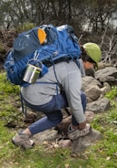 A person kneels on rocky terrain surrounded by greenery, adjusting their hiking boots. They are equipped with a large blue backpack that includes various outdoor gear such as a metal mug and a rolled-up mat. The person is wearing a green cap, a gray long-sleeve top, and blue pants.
