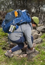 A person kneels on rocky terrain surrounded by greenery, adjusting their hiking boots. They are equipped with a large blue backpack that includes various outdoor gear such as a metal mug and a rolled-up mat. The person is wearing a green cap, a gray long-sleeve top, and blue pants.