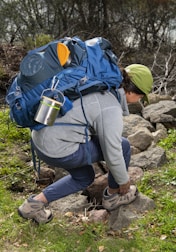 A person kneels on rocky terrain surrounded by greenery, adjusting their hiking boots. They are equipped with a large blue backpack that includes various outdoor gear such as a metal mug and a rolled-up mat. The person is wearing a green cap, a gray long-sleeve top, and blue pants.