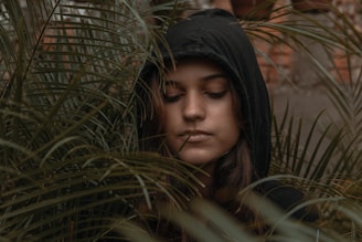 A person wearing a dark hooded sweatshirt stands amidst dense green foliage with eyes closed, expressing a serene or contemplative mood. The leaves surround their face, creating an intimate, natural setting.
