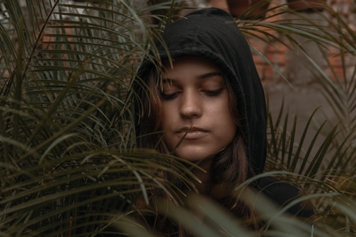 A person wearing a dark hooded sweatshirt stands amidst dense green foliage with eyes closed, expressing a serene or contemplative mood. The leaves surround their face, creating an intimate, natural setting.