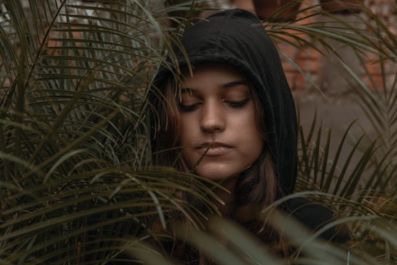 A person wearing a dark hooded sweatshirt stands amidst dense green foliage with eyes closed, expressing a serene or contemplative mood. The leaves surround their face, creating an intimate, natural setting.