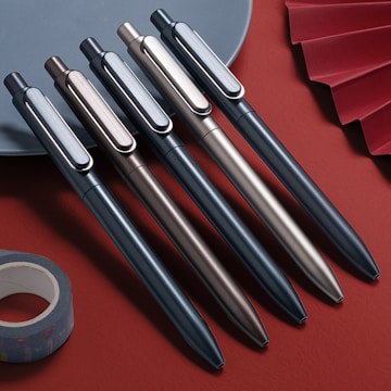 A neatly arranged row of five sleek metal pens in varying metallic shades, placed atop a red surface. The pens are aligned parallel to each other, with the click mechanism and pocket clip visible. A roll of decorative tape and a hint of a folded paper fan are also present, adding to the artistic composition.