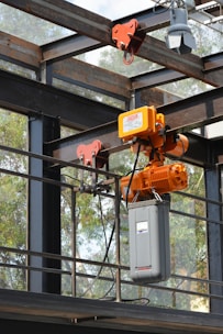 A heavy-duty industrial crane mechanism is attached to a metal structure, with orange machinery and a large gray casing. The mechanism is situated outdoors, surrounded by a metal frame and glass panels. Trees can be seen in the background through the glass.