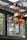 A heavy-duty industrial crane mechanism is attached to a metal structure, with orange machinery and a large gray casing. The mechanism is situated outdoors, surrounded by a metal frame and glass panels. Trees can be seen in the background through the glass.