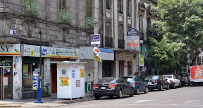 A street scene with a row of parked cars in front of various storefronts. The buildings are older with intricate architectural details, including wrought iron railings on the balconies. Signs in Spanish advertise different businesses, including a pharmacy and a restaurant. A tree is visible, providing some greenery to the urban setting.