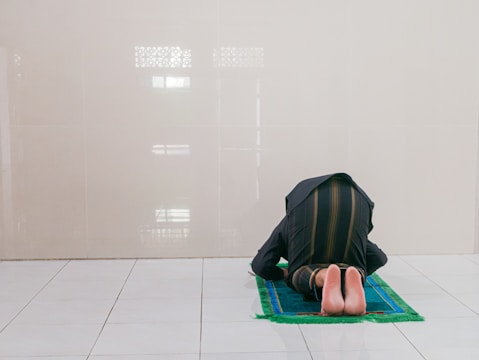 A person is in a kneeling position on a prayer mat, facing a tiled wall. The mat has a green border and a blue center. The space is minimal with a reflective floor and muted wall colors.