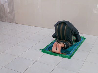 A person is kneeling on a blue and green prayer mat in a tiled room, with their head resting on the mat and feet visible from behind. The person's robe covers their head and body.