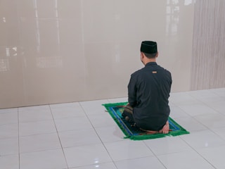 A person is sitting on a prayer mat facing a blank wall in a tiled room. The individual is dressed in a black outfit including a cap. The overall environment appears to be clean and minimalistic.