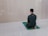 A person is sitting on a prayer mat facing a blank wall in a tiled room. The individual is dressed in a black outfit including a cap. The overall environment appears to be clean and minimalistic.
