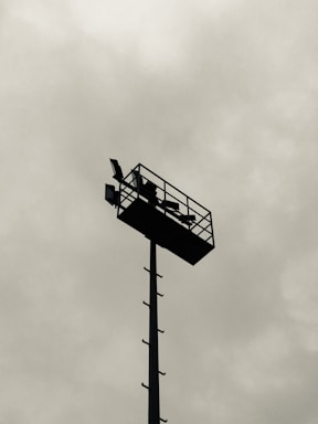 A tall metal pole with a rectangular platform at the top, featuring mounted lights and a ladder along the side, set against a cloudy sky.