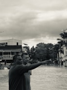 Two individuals stand amidst what appears to be a flood, with one person pointing towards something in the distance. Surrounding them are partially submerged buildings and trees, under a cloudy sky. The setting suggests an urban area experiencing heavy flooding.