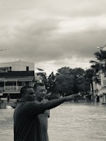 Two individuals stand amidst what appears to be a flood, with one person pointing towards something in the distance. Surrounding them are partially submerged buildings and trees, under a cloudy sky. The setting suggests an urban area experiencing heavy flooding.