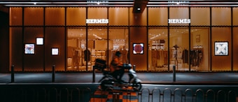 A sleek display of a Hermes store with illuminated signage and stylish window designs. A person on a scooter rides past the store, creating a dynamic contrast with the stationary elegance of the storefront.