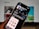 A smartphone displays the Clubhouse app in the Apple App Store. The app's page shows information such as its name, ratings, and age suitability. The background is blurred, showing a computer screen.
