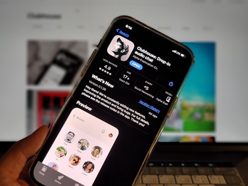 A smartphone displays the Clubhouse app in the Apple App Store. The app's page shows information such as its name, ratings, and age suitability. The background is blurred, showing a computer screen.