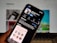A smartphone displays the Clubhouse app in the Apple App Store. The app's page shows information such as its name, ratings, and age suitability. The background is blurred, showing a computer screen.