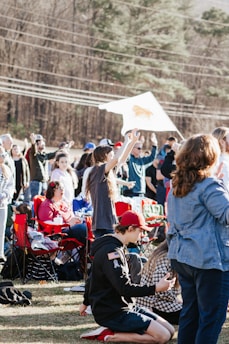 A group of people gathered outdoors, some sitting on folding chairs and others standing or kneeling. They appear to be involved in a public or community event. A large white flag with a design is visible in the background, and the setting is surrounded by trees and power lines.