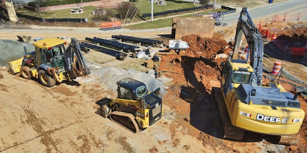 A construction site is depicted with heavy machinery including an excavator, a bulldozer, and a backhoe loader. Large pipes are stacked in the background, and there is a section of earth that appears to be excavated. Traffic cones and barriers are positioned around the site, indicating ongoing work. The ground is mostly bare and dusty, with some areas containing gravel and red dirt.