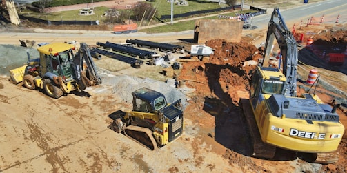 A construction site is depicted with heavy machinery including an excavator, a bulldozer, and a backhoe loader. Large pipes are stacked in the background, and there is a section of earth that appears to be excavated. Traffic cones and barriers are positioned around the site, indicating ongoing work. The ground is mostly bare and dusty, with some areas containing gravel and red dirt.