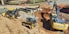 A construction site is depicted with heavy machinery including an excavator, a bulldozer, and a backhoe loader. Large pipes are stacked in the background, and there is a section of earth that appears to be excavated. Traffic cones and barriers are positioned around the site, indicating ongoing work. The ground is mostly bare and dusty, with some areas containing gravel and red dirt.