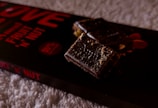 A close-up view of a chocolate bar with nuts, placed on its packaging. The chocolate appears rich and dark with visible nut pieces, and the packaging displays bold red text indicating 'fruit 'n' nut' and '55% cocoa'. The background is a soft, textured surface.
