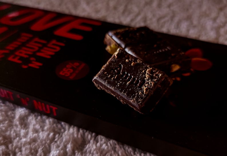 A close-up view of a chocolate bar with nuts, placed on its packaging. The chocolate appears rich and dark with visible nut pieces, and the packaging displays bold red text indicating 'fruit 'n' nut' and '55% cocoa'. The background is a soft, textured surface.