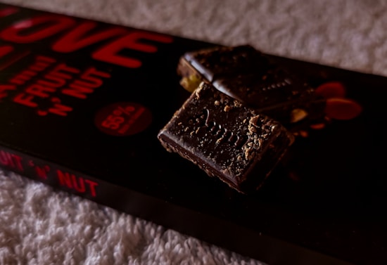 A close-up view of a chocolate bar with nuts, placed on its packaging. The chocolate appears rich and dark with visible nut pieces, and the packaging displays bold red text indicating 'fruit 'n' nut' and '55% cocoa'. The background is a soft, textured surface.
