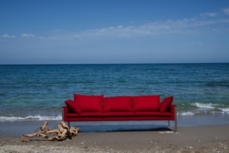 A bright red sofa is placed on a sandy beach with the ocean in the background. Gentle waves approach the shore under a clear blue sky. A piece of driftwood lies on the sand near the sofa.
