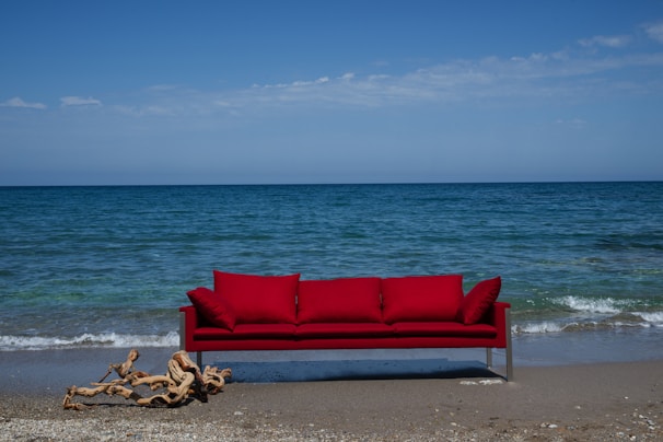 A bright red sofa is placed on a sandy beach with the ocean in the background. Gentle waves approach the shore under a clear blue sky. A piece of driftwood lies on the sand near the sofa.