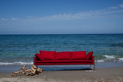 A bright red sofa is placed on a sandy beach with the ocean in the background. Gentle waves approach the shore under a clear blue sky. A piece of driftwood lies on the sand near the sofa.
